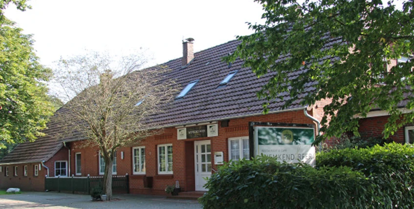 Außenansicht Ein rotes Backsteingebäude mit vielen Fenstern unter einem Schrägdach, umgeben von Bäumen.