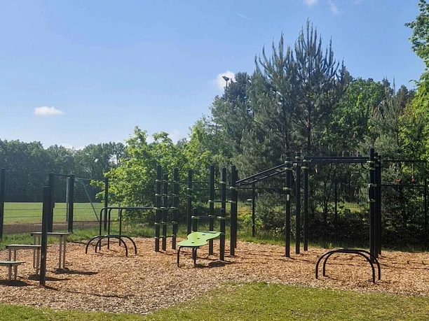 Bild-Outdoorfitnessanlage-Groß-Hehlen-Hochformat.jpg Outdoor-Fitnessanlage mit diversen Sportgeräten auf einem Holzhackschnitzel-Boden vor Bäumen.