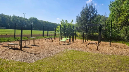 Outdoorfitness-Anlage Groß Hehlen Verschiedene Fitnessgeräte auf einer Holzschnitzel-Fläche, umgeben von Bäumen und einer Wiese.