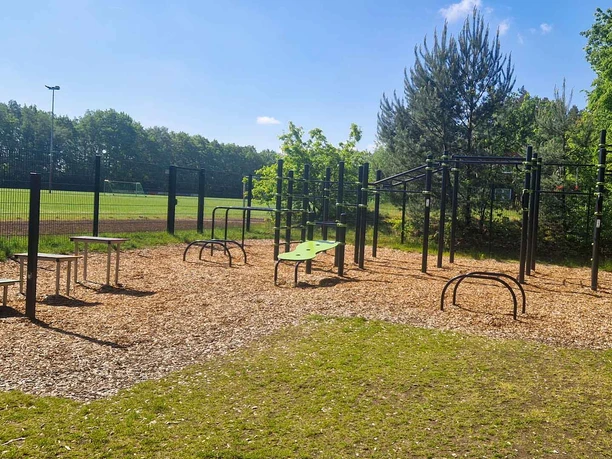 Outdoorfitness-Anlage Groß Hehlen Verschiedene Fitnessgeräte auf einer Holzschnitzel-Fläche, umgeben von Bäumen und einer Wiese.
