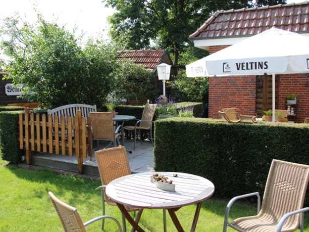 Terrasse2 Ein gepflegter Garten mit Terrasse. Im Vordergrund ein runder Holztisch mit zwei Stühlen. Im Hintergrund stehen weitere Sitzgelegenheiten und ein Sonnenschirm, der Schatten spendet. Umgeben von grünen Hecken und Bäumen, lädt die Umgebung zum Entspannen ein.