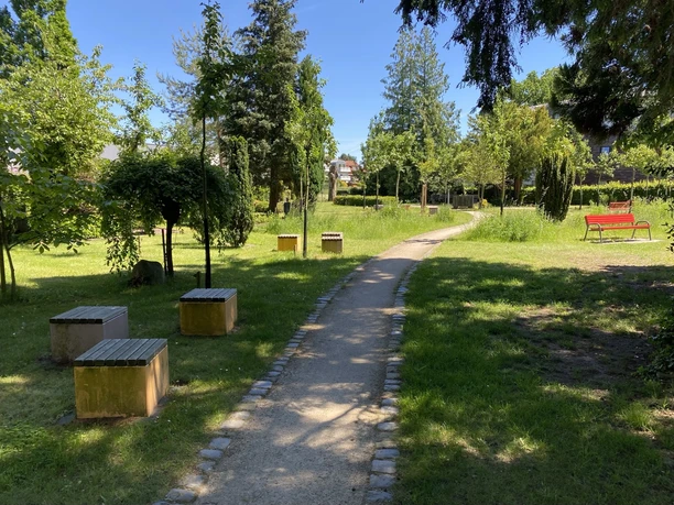 Apfelgärtchen Alter Friedhof Gemeinde Steinhagen Geschwungener Weg durch Apfelgarten mit Bäumen, Sitzwürfeln und roter Bank im Park.