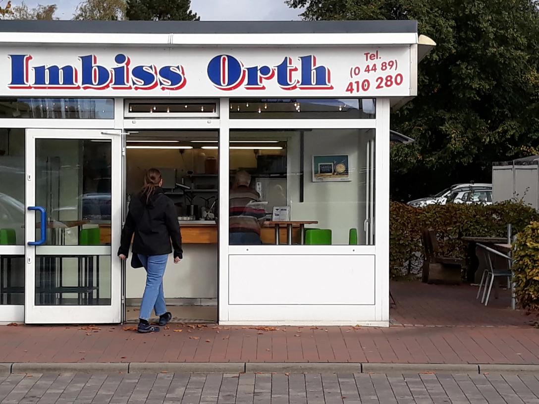 Imbiss Orth Das Bild zeigt einen kleinen, weißen Imbissstand mit dem Namen "Imbiss Orth". Eine Person nähert sich dem Eingang.