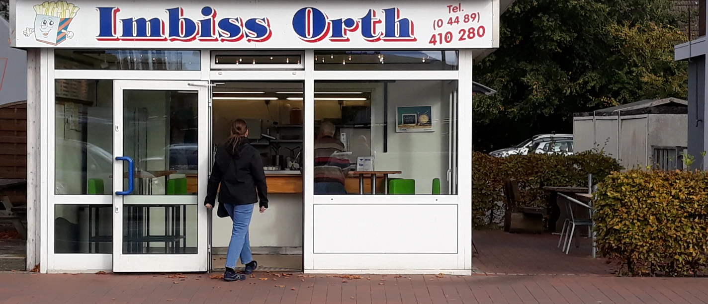 Imbiss Orth Das Bild zeigt einen kleinen, weißen Imbissstand mit dem Namen "Imbiss Orth". Eine Person nähert sich dem Eingang.