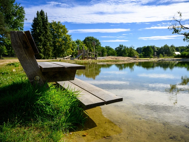 DK_25_112496.jpg Holzbank am Ufer des Badeteichs Steinbeck mit Spielplatz und Sandstrand im Hintergrund.