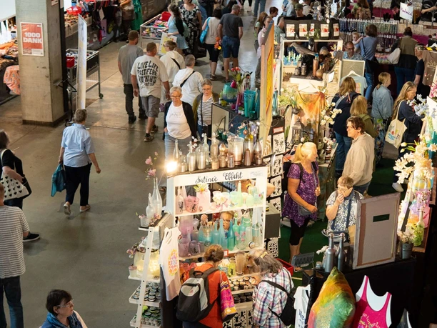 Kölner Creativ Sommer Menschen schlendern durch eine Messehalle mit dicht gereihten Kreativständen und vielfältigen Produkten.