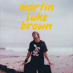 Person am Strand mit dunklem Shirt und gelber Schrift martin luke brown darüber
