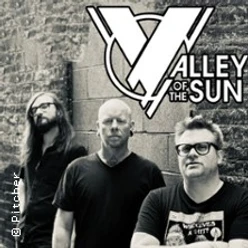 Drei Musiker stehen vor einer Backsteinwand, darüber das Valley‑of‑the‑Sun‑Logo im Grafikstil.