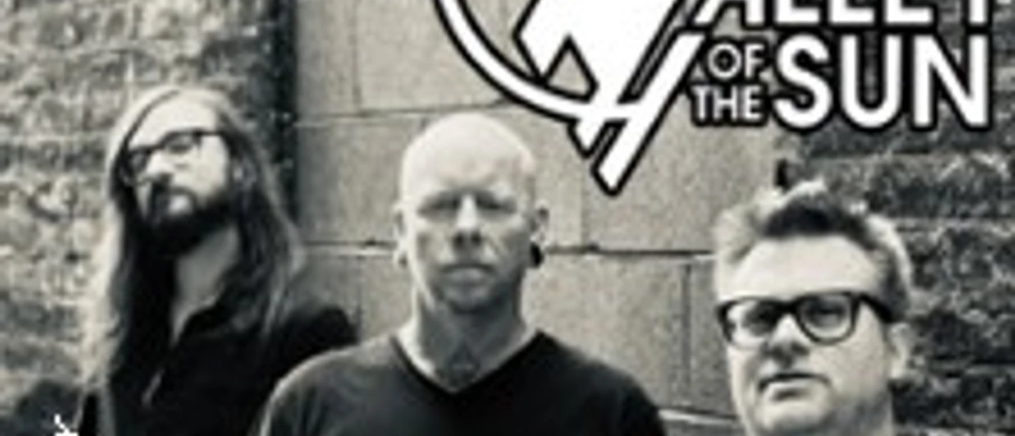 Drei Musiker stehen vor einer Backsteinwand, darüber das Valley‑of‑the‑Sun‑Logo im Grafikstil.