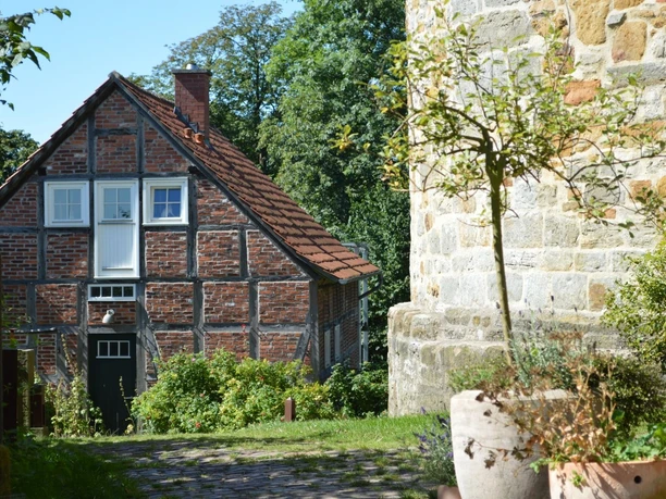 Burg Ravensberg, Borgholzhausen - Grünes Klassenzimmer Fachwerkhaus neben einer steinernen Burgmauer, umgeben von Pflanzen und unter blauem Himmel.