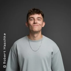 Junger Mann in hellem Shirt vor dunklem Hintergrund mit entspanntem Gesichtsausdruck.