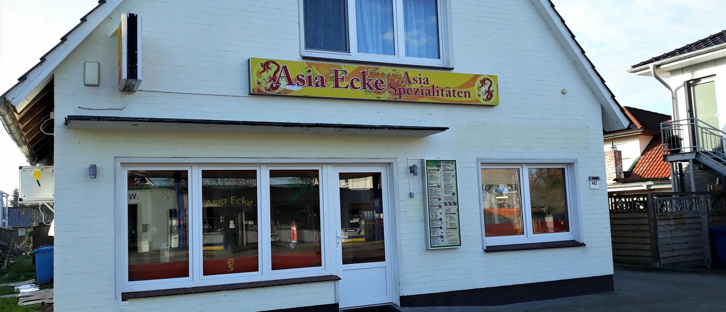 Asia Ecke Das Bild zeigt ein kleines, weißes Gebäude mit einem Giebel, das ein asiatisches Restaurant beherbergt. Über dem Eingang hängt ein Schild mit der Aufschrift „Asia Ecke – Asia Spezialitäten“, flankiert von roten Drachenmotiven. Große Fenster in der Front verheißen ein helles Inneres, während das Kopfsteinpflaster den rustikalen Charme des Ortes unterstreicht.