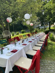 Sundown Dinner Langer Gartentisch mit weißer Tafel, bunten Stühlen und Laternen unter Bäumen.
