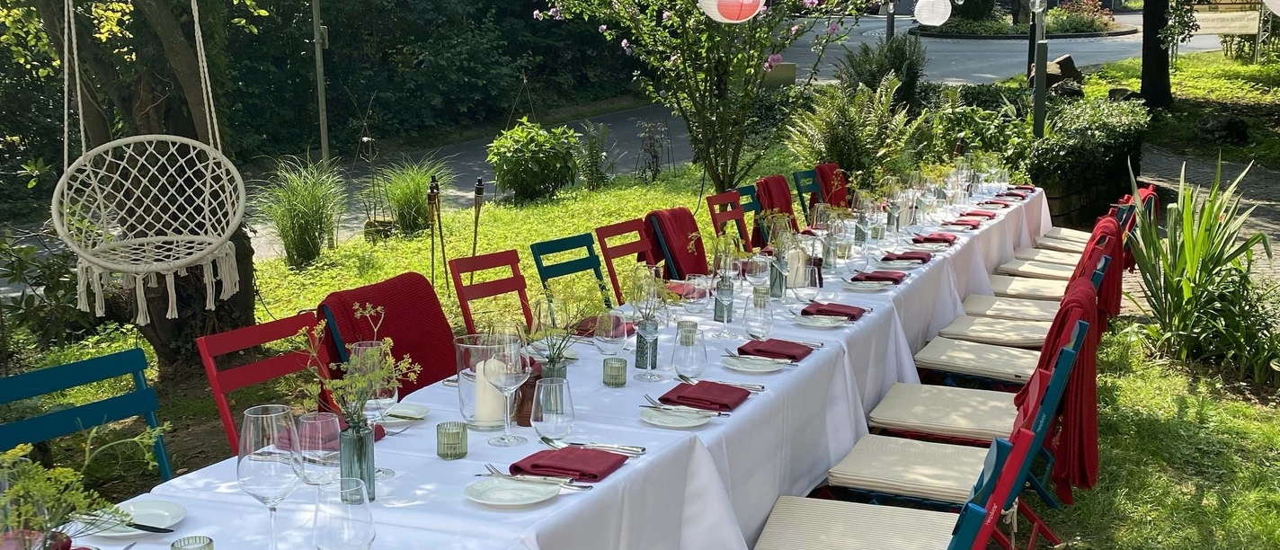 Sundown Dinner Langer Gartentisch mit weißer Tafel, bunten Stühlen und Laternen unter Bäumen.