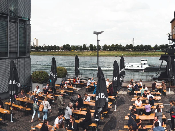 Belebter Biergarten am Rhein mit Holzbänken, Sonnenschirmen und altem Hafenkran.