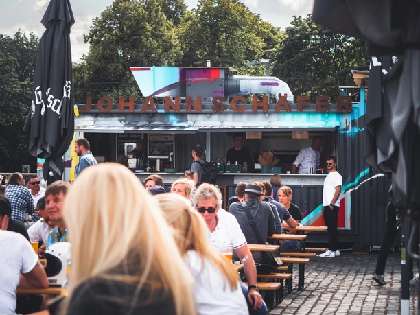 Johann Schäfer Biergarten am Rhein im Rheinauhafen Lebhafter Biergarten mit Containerbar und vielen Gästen an Holzbänken unter freiem Himmel