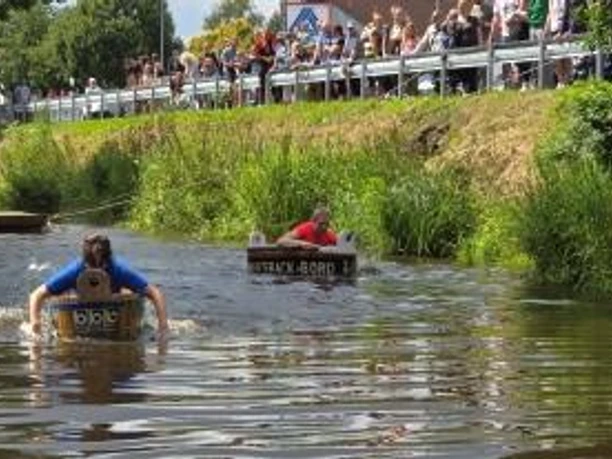 Waschzuber Regatta.jpg Teilnehmer eines Wettrennens paddeln in umgebauten Fässern auf einem Kanal vor Publikum am Ufer.