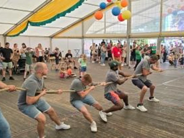 Firmen Nachmittag.jpg Männer ziehen beim Tauziehen in einem Festzelt kräftig an einem Seil, angefeuert von Zuschauern.