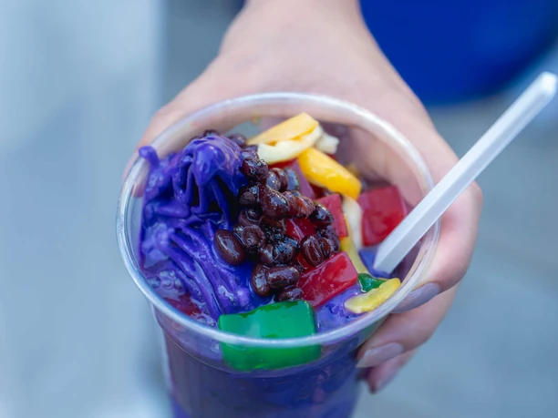 Ube Fiesta Hand hält einen Becher mit violettem Dessert und bunten Frucht- und Gelee-Stücken.
