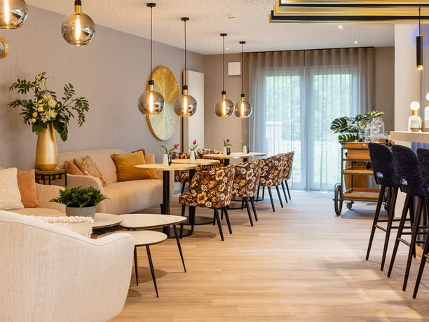 Claras Restaurant (2) Moderner Lounge‑ und Barbereich im Claras Restaurant mit floralen Polstersesseln, hellen Sofas, gläsernen Hängeleuchten und einer Bartheke mit hohen blauen Hockern.
