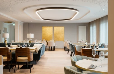 Claras Restaurant Wolfsburg.png Heller, elegant eingerichteter Gastraum im Claras Restaurant mit gedeckten Tischen, bequemen Polsterstühlen und zwei auffälligen goldfarbenen Kunsttafeln an der Rückwand.
