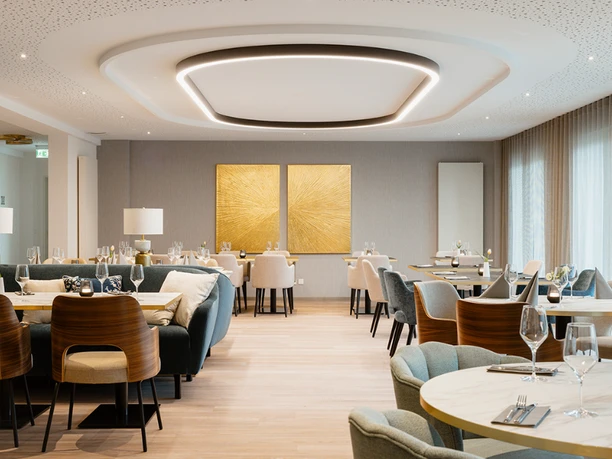 Claras Restaurant Wolfsburg.png Heller, elegant eingerichteter Gastraum im Claras Restaurant mit gedeckten Tischen, bequemen Polsterstühlen und zwei auffälligen goldfarbenen Kunsttafeln an der Rückwand.