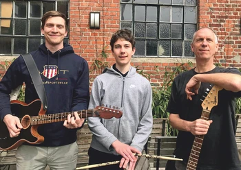 Drei Musiker des Mark Scheel Trios mit Gitarren und Schlagzeugsticks vor Backsteinwand
