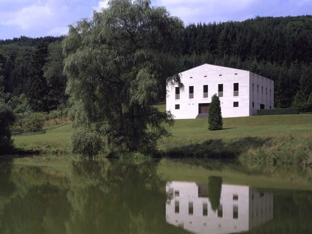O.M. Ungers, Haus Glashütte, Eifel 1986-1988 Bright geometric building by a pond with a large pasture and wooded slope in the background