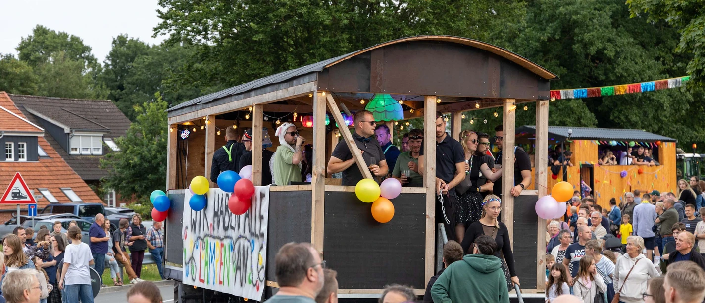 Festwagen.jpg Ein geschmückter Festwagen mit feiernden Menschen fährt bei einem Umzug an Zuschauern vorbei.