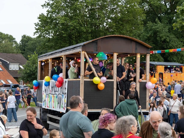 Festwagen.jpg Ein geschmückter Festwagen mit feiernden Menschen fährt bei einem Umzug an Zuschauern vorbei.
