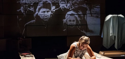 Bühnenszene zu Anne Frank: Darstellerin schreibt vor projizierten Kinderporträts.