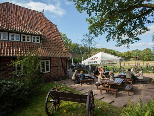 gasthaus-zum-heidemuseum Das Bild zeigt ein traditionelles, reetgedecktes Gasthaus aus rotem Backstein. Vor dem Gebäude sitzen Gäste an Holztischen im Freien unter einem weißen Sonnenschirm. Links im Vordergrund befindet sich ein alter Holzkarren auf einer Rasenfläche. Im Hintergrund erstreckt sich eine Wiese.