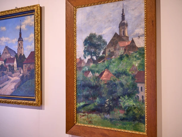 Zwei gerahmte Landschaftsgemälde an einer Wand, eines mit Kirchen und Häusern, das andere mit grünen Hügeln.