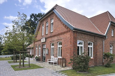 Gristeder Hof Das Bild zeigt ein hübsches, großes Gebäude aus rotem Backstein mit modernen weißen Fensterrahmen. Es ist der Gristeder Hof, ein traditionelles, zweigeschossiges Haus mit einem steilen Ziegeldach. Vor dem Gebäude läuft eine gepflasterte Fläche entlang, auf der sich Fahrradbügel und eine Bank befinden. Ein kleiner Baum steht am linken Rand. Im Hintergrund ist Himmel mit vereinzelten Wolken zu sehen.