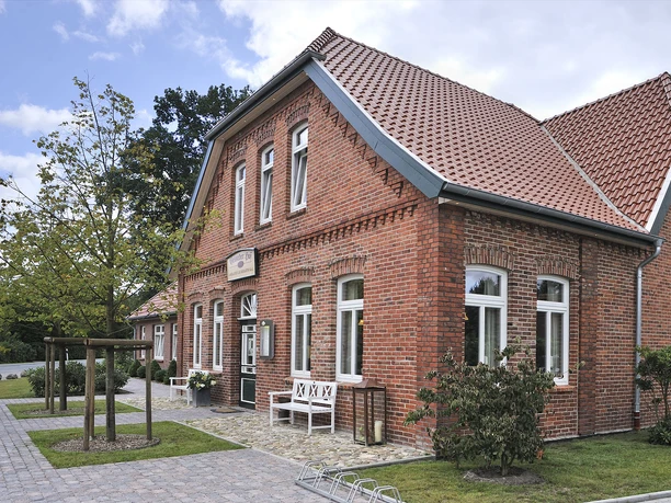 Gristeder Hof Das Bild zeigt ein hübsches, großes Gebäude aus rotem Backstein mit modernen weißen Fensterrahmen. Es ist der Gristeder Hof, ein traditionelles, zweigeschossiges Haus mit einem steilen Ziegeldach. Vor dem Gebäude läuft eine gepflasterte Fläche entlang, auf der sich Fahrradbügel und eine Bank befinden. Ein kleiner Baum steht am linken Rand. Im Hintergrund ist Himmel mit vereinzelten Wolken zu sehen.