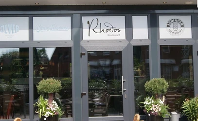 Rhodos Das Bild zeigt den Eingang des Restaurants "Rhodos". Es hat große Glasfenster und einen grauen Rahmen. Vor der Tür stehen zwei grüne Pflanzen.