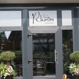 Rhodos Das Bild zeigt den Eingang des Restaurants "Rhodos". Es hat große Glasfenster und einen grauen Rahmen. Vor der Tür stehen zwei grüne Pflanzen.
