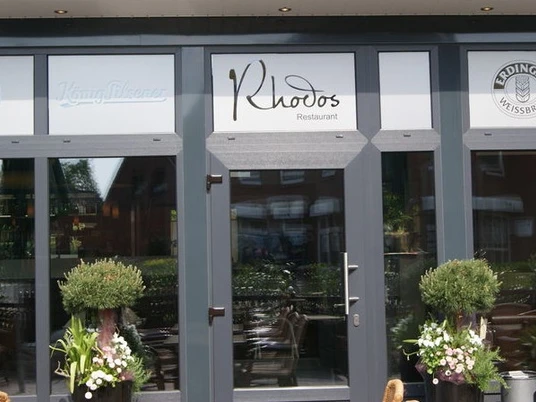 Rhodos Das Bild zeigt den Eingang des Restaurants "Rhodos". Es hat große Glasfenster und einen grauen Rahmen. Vor der Tür stehen zwei grüne Pflanzen.