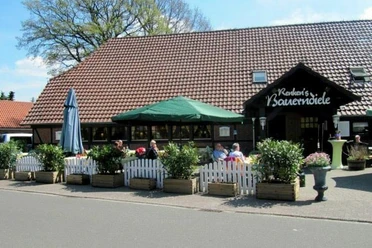 Renkens Das Bild zeigt ein gemütliches Café mit roten Ziegeldach, umgeben von Pflanzen in Holzkübeln.