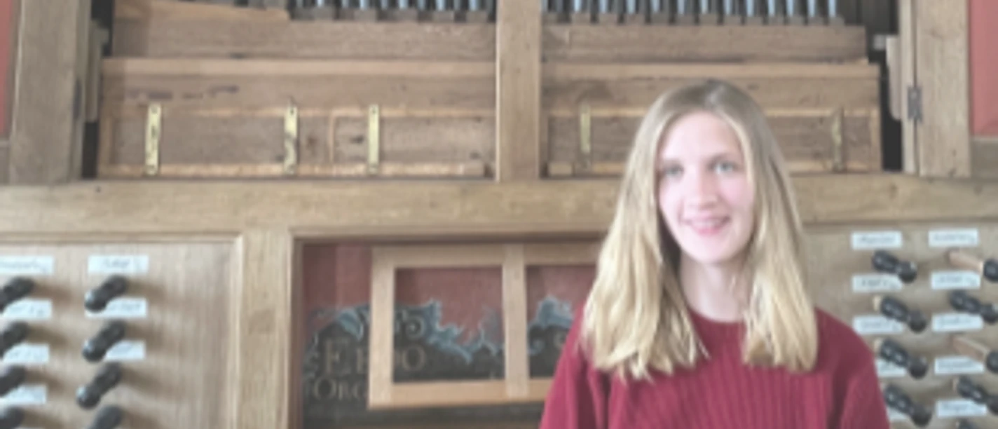 Musikalische Andacht mit Orgelmusik Junge Frau in rotem Pullover lächelt vor der großen, hölzernen Pfeifenorgel in der Pilsumer Kirche.