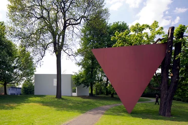 Parkansicht Skulpturenpark Köln Roter invertierter Dreieckskörper im Park mit Bäumen, Weg und modernem hellen Bau.