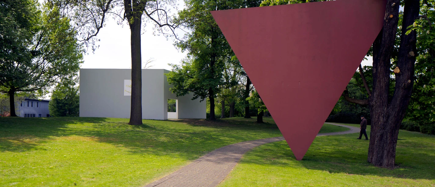 Parkansicht Skulpturenpark Köln Roter invertierter Dreieckskörper im Park mit Bäumen, Weg und modernem hellen Bau.