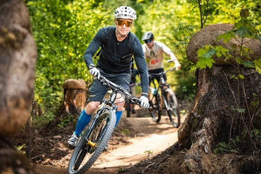 Borderlesstrails.jpg Zwei Mountainbiker fahren auf einem schmalen Waldtrail durch eine naturnahe Waldlandschaft. Im Vordergrund fährt ein Radfahrer durch eine Kurve, während ein weiterer Fahrer ihm auf dem Trail folgt.