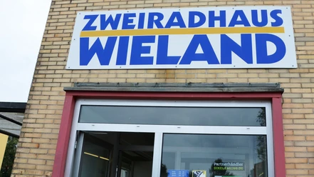 Wieland_Fahrradhandel (5)_HD_16zu9.JPG Der Eingang des Zweiradhaus Wieland mit großem Firmenschild über der Tür an einer Backsteinfassade.