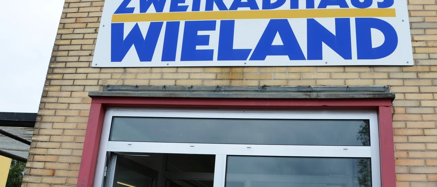 Wieland_Fahrradhandel (5)_HD_16zu9.JPG Der Eingang des Zweiradhaus Wieland mit großem Firmenschild über der Tür an einer Backsteinfassade.