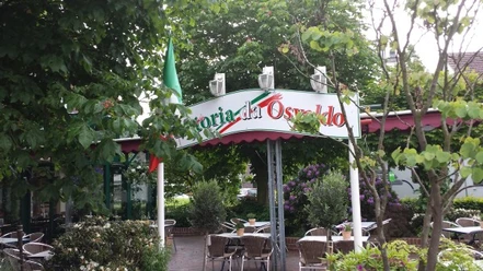trattoria_osvaldo Auf dem Bild ist der Eingang der Trattoria Osvaldo zu sehen. Ein Bogenschild zeigt den Namen der Trattoria an. Der Bereich ist von grünen Bäumen umgeben und es gibt Sitzgelegenheiten im Freien.