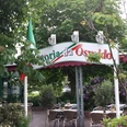 trattoria_osvaldo Auf dem Bild ist der Eingang der Trattoria Osvaldo zu sehen. Ein Bogenschild zeigt den Namen der Trattoria an. Der Bereich ist von grünen Bäumen umgeben und es gibt Sitzgelegenheiten im Freien.