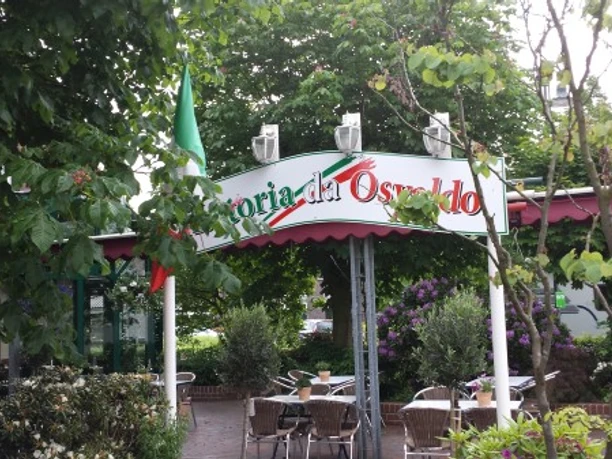 trattoria_osvaldo Auf dem Bild ist der Eingang der Trattoria Osvaldo zu sehen. Ein Bogenschild zeigt den Namen der Trattoria an. Der Bereich ist von grünen Bäumen umgeben und es gibt Sitzgelegenheiten im Freien.