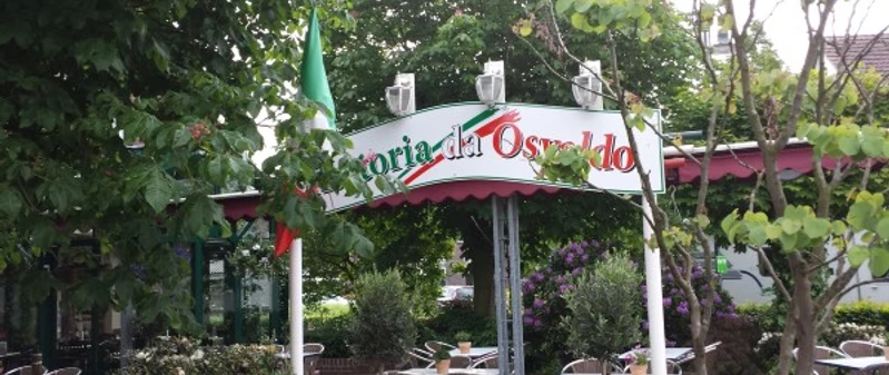 trattoria_osvaldo Auf dem Bild ist der Eingang der Trattoria Osvaldo zu sehen. Ein Bogenschild zeigt den Namen der Trattoria an. Der Bereich ist von grünen Bäumen umgeben und es gibt Sitzgelegenheiten im Freien.