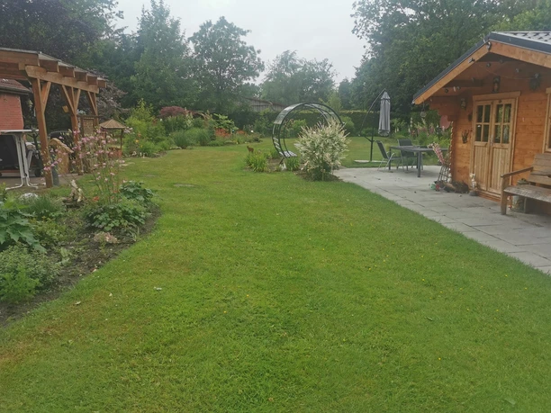 Blockhaus mit Sicht in den Garten_Foto Hienen.jpg Große Rasenfläche in einem Garten, gesäumt von Beeten, mit einem Holzhaus und Terrasse rechts.
